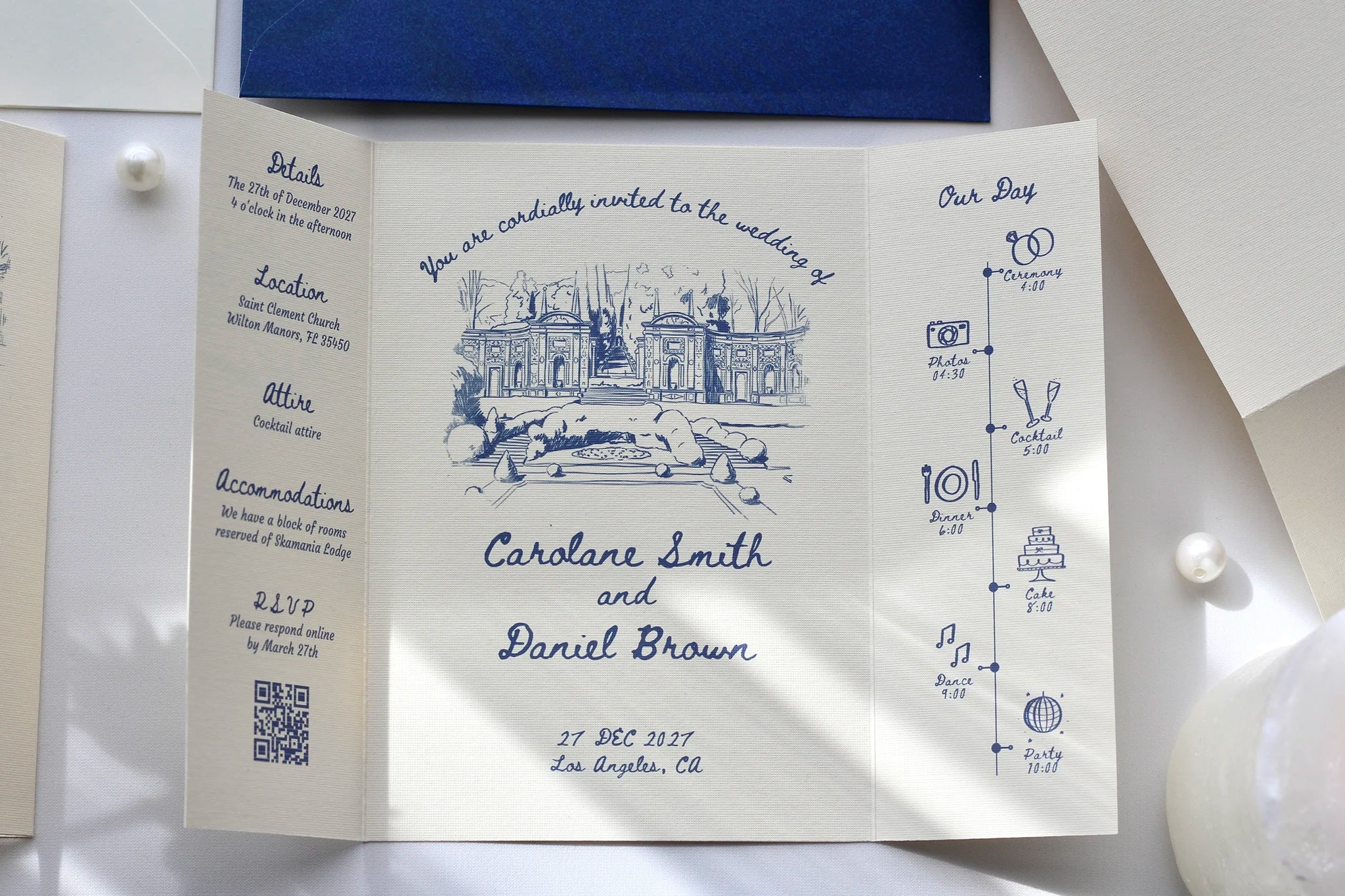 A white flat-lay wedding invitation card with blue ink illustrations and text, displayed on a white surface with a dark blue fabric and pearl-like beads nearby.