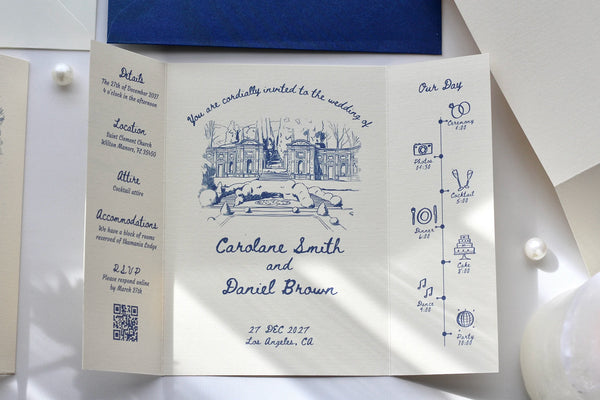 A white flat-lay wedding invitation card with blue ink illustrations and text, displayed on a white surface with a dark blue fabric and pearl-like beads nearby.