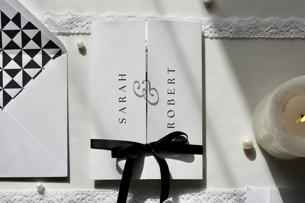A white wedding invitation card laid flat on a lace-trim surface, surrounded by black-and-white stationery and accessories. The card features elegant script text reading 'Sarah & Robert' and event details.