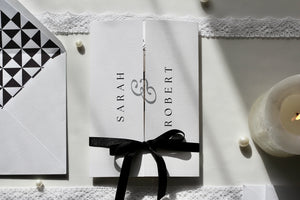 A white wedding invitation card set with a black ribbon bow, featuring the names 'SARAH' and 'ROBERT' with a decorative monogram. Styled flat-lay with a black-and-white geometric envelope, lace trim, pearls, and a lit candle.