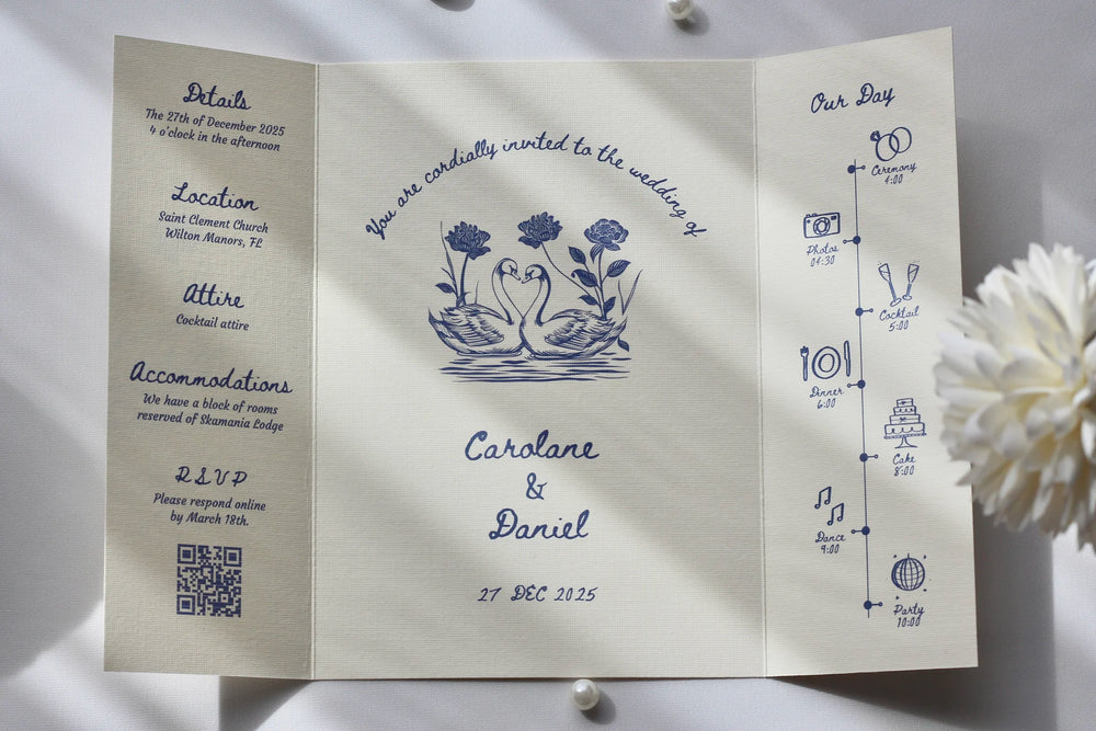 A wedding invitation suite displayed on a light gray surface with decorative white flowers and shadows. The set includes a folded invitation card with blue floral and timeline illustrations, a matching folded card showing event details, and a separate card with event text.