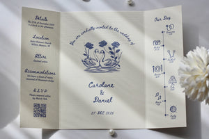 A folded white wedding invitation card laid flat on a white fabric surface with pearl beads and a white flower prop. The card has navy blue printed text and illustrations, including a central floral motif and event details for Caroline & Daniel.