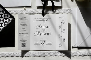 A white wedding invitation card laid flat on a lace-trim surface, surrounded by black-and-white stationery and accessories. The card features elegant script text reading 'Sarah & Robert' and event details.
