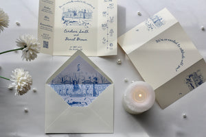 A styled flat-lay of a wedding invitation suite on a light gray surface, featuring cream-colored paper cards and envelopes with blue architectural line drawings and elegant script text. White flowers, small pearl beads, and a lit white candle are arranged around the stationery.