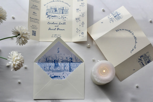 A styled flat-lay of a wedding invitation suite on a light gray surface, featuring cream-colored paper cards and envelopes with blue architectural line drawings and elegant script text. White flowers, small pearl beads, and a lit white candle are arranged around the stationery.