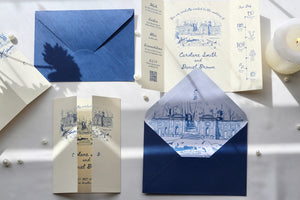 A styled flat-lay of blue and cream stationery, including an envelope, invitation cards, and a folded card with blue architectural sketches on cream paper, arranged on a light surface with shadows and small pearl-like beads.