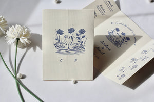 A cream-colored stationery set featuring a blue ink illustration of two swans under four flowers, displayed on a white surface with white flowers and soft shadows.