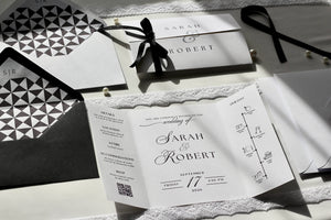 A styled flat-lay of wedding stationery including a white invitation card with elegant black typography, a matching envelope, a black ribbon bow, and geometric patterned paper pieces on a light surface.