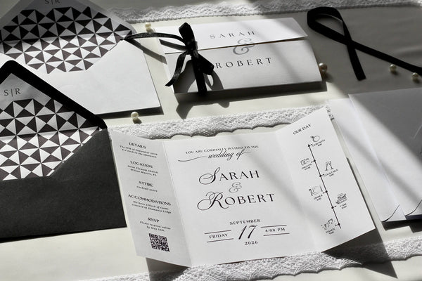 A styled flat-lay of wedding stationery including a white invitation card with elegant black typography, a matching envelope, a black ribbon bow, and geometric patterned paper pieces on a light surface.