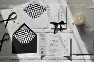 A styled flat-lay of wedding stationery on a white lace-trimmed surface, including a white invitation card with black script text, matching envelopes, a black-and-white geometric patterned insert, black ribbons, and a small lit candle.