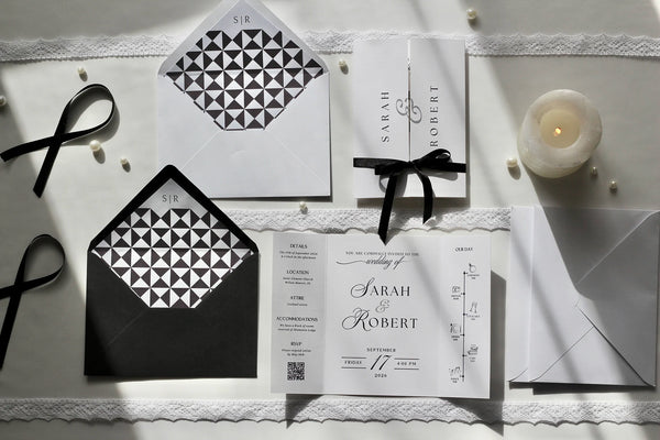 A styled flat-lay of wedding stationery on a white lace-trimmed surface, including a white invitation card with black script text, matching envelopes, a black-and-white geometric patterned insert, black ribbons, and a small lit candle.