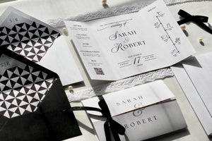 A styled flat-lay of wedding stationery featuring a white gatefold invitation with elegant black typography, a matching envelope with a black and white geometric pattern, and coordinating cards tied with black ribbons on a light fabric background.