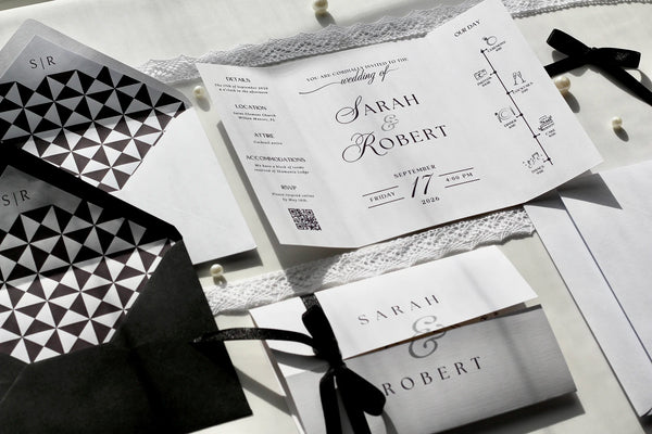 A styled flat-lay of wedding stationery featuring a white gatefold invitation with elegant black typography, a matching envelope with a black and white geometric pattern, and coordinating cards tied with black ribbons on a light fabric background.
