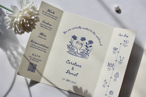 An open tri-fold wedding invitation card displayed on a light surface with a white flower and pearl nearby. The card has a warm cream background with dark blue printed text and illustrations, including a central swan motif and timeline details.