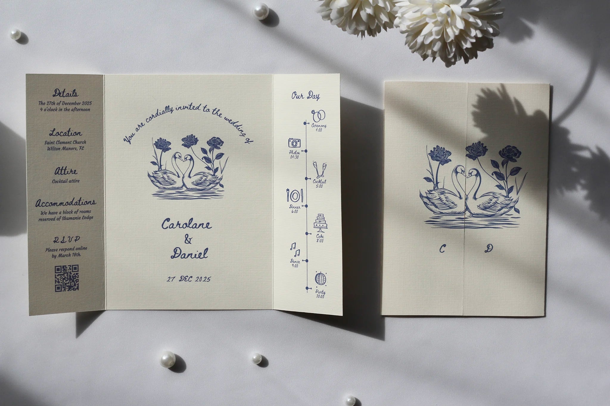 A wedding invitation suite displayed on a light gray surface with decorative white flowers and shadows. The set includes a folded invitation card with blue floral and timeline illustrations, a matching folded card showing event details, and a separate card with event text.