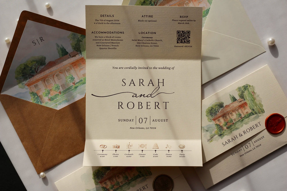 A styled flat-lay of wedding stationery, featuring a central cream invitation card with black typography and a watercolor villa illustration, surrounded by a brown envelope, a matching printed card with a red wax seal, and other wedding details on a white surface.