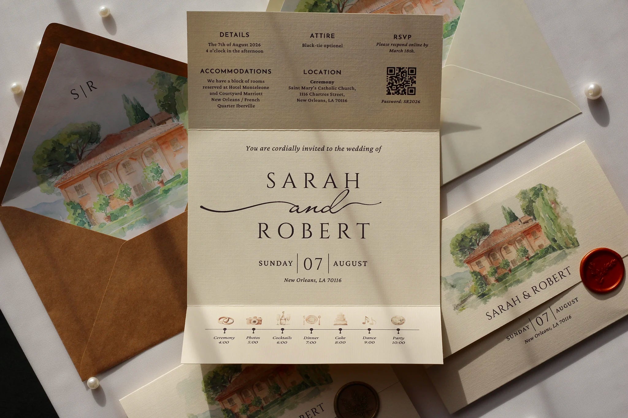 A styled flat-lay of wedding stationery, featuring a central cream invitation card with black typography and a watercolor villa illustration, surrounded by a brown envelope, a matching printed card with a red wax seal, and other wedding details on a white surface.