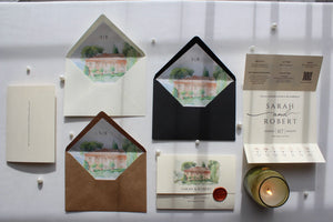 A coordinated stationery set arranged on a white surface, including cream and kraft envelopes with watercolor architectural liner cards, a printed card, a small candle, and a printed insert. No person, animal, or real environment is present, so this is Product/Packaging rather than Lifestyle.