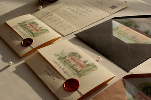 A styled flat-lay of elegant stationery featuring cream invitation cards with watercolor illustrations of a villa, a dark gray envelope, and red wax seals on a neutral surface.