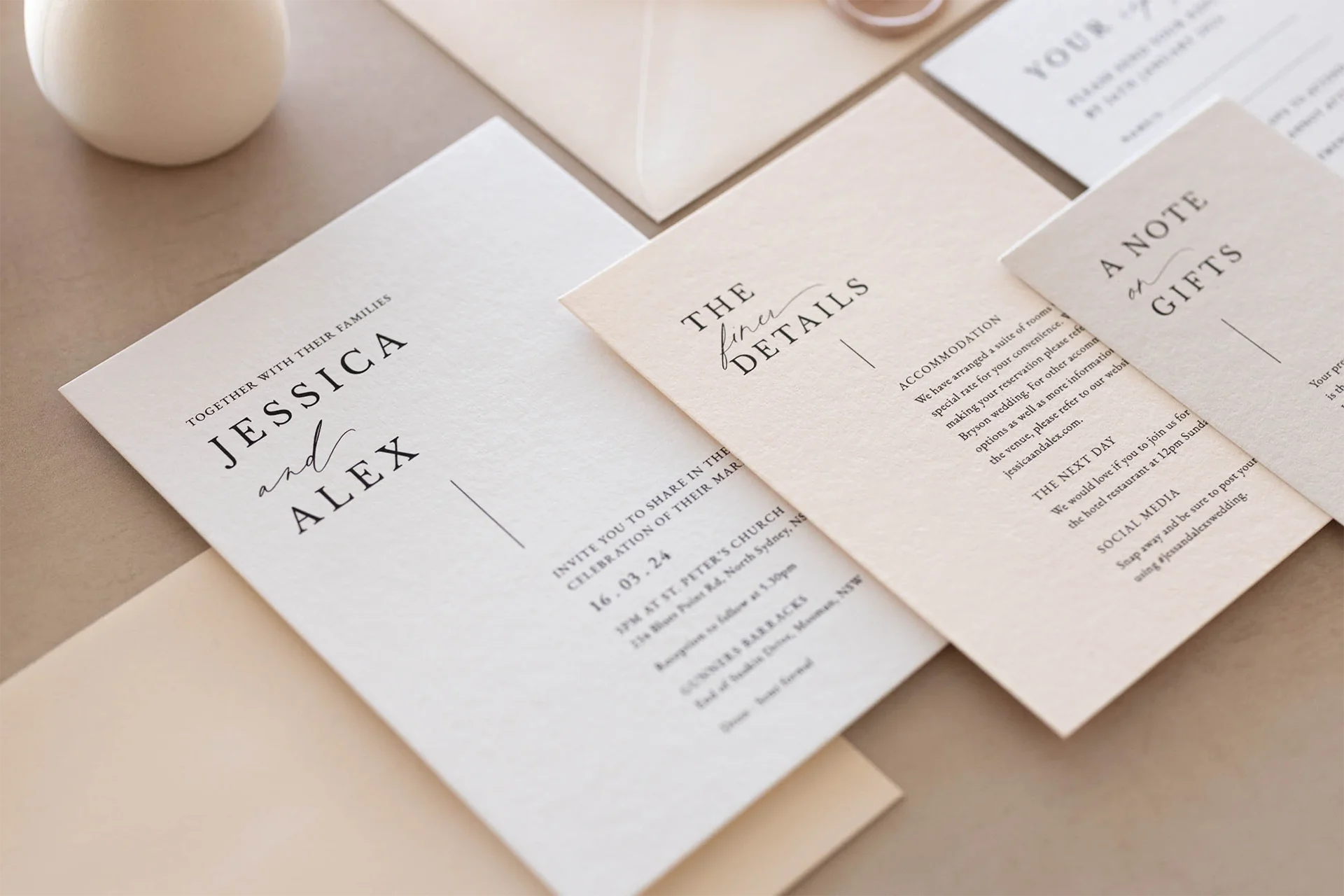 A styled flat-lay of a wedding invitation suite featuring cream and off-white paper cards with elegant black typography, arranged on a neutral beige surface with a small white decorative object.