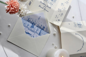 A styled flat-lay of wedding stationery on a light surface, including an open cream envelope with a blue architectural illustration liner, a printed invitation card, a folded card, small pearl-like beads, flowers, and a lit candle.